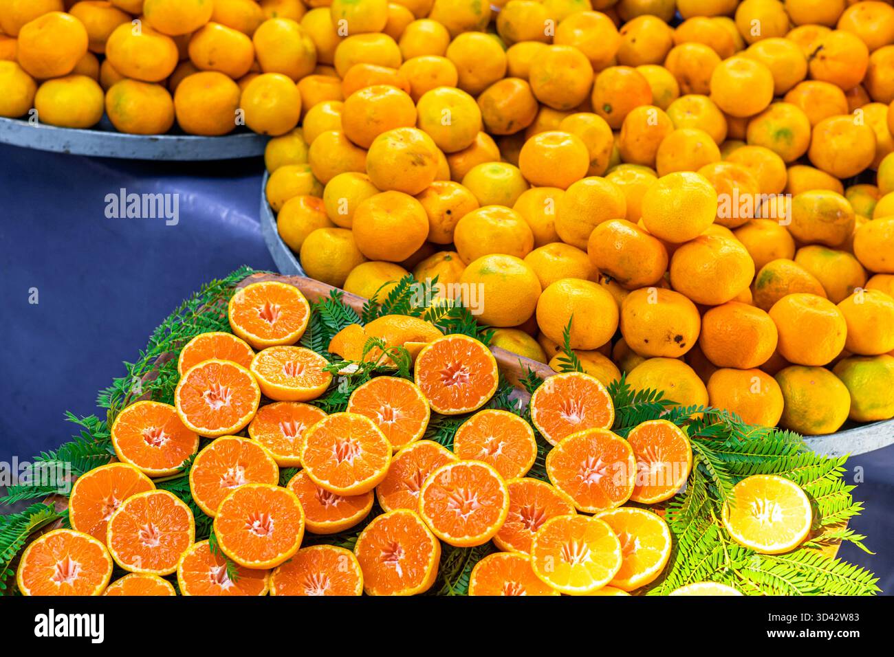 Frische Orangen und Mandarinen auf einem Markt. Ganze Orangen sind hoch gestapelt, während geschnittene Orangen auf einem Bett aus grünen Blättern angeordnet sind. Stockfoto