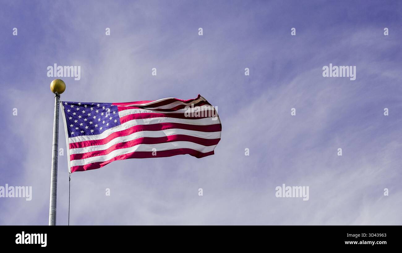 Die amerikanische Flagge fliegt stolz auf einem Fahnenmast und flattert sanft in der Brise unter einem klaren blauen Himmel. Helles Sonnenlicht beleuchtet seine leuchtenden Farben. Stockfoto