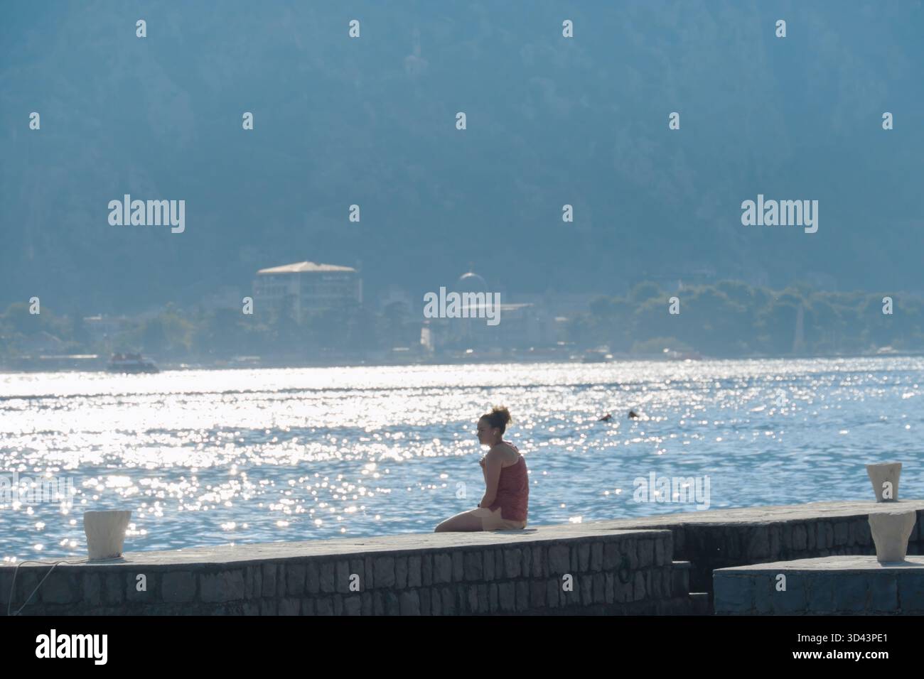 Junge Frau, die am Ufer sitzt und sich bei Morgensonne in der Bucht von Kotor in Montenegro entspannt Musik hört Stockfoto