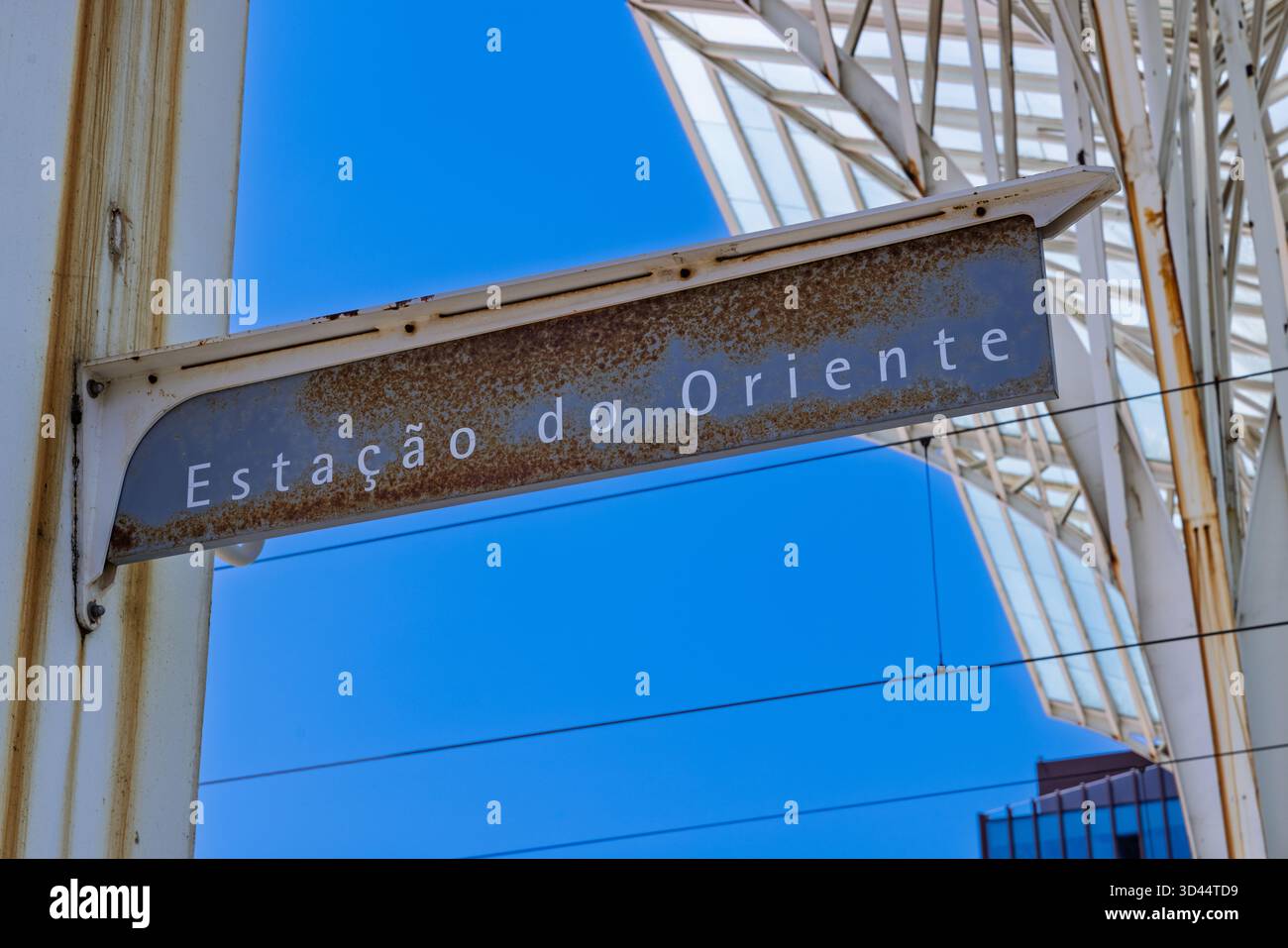 Nahaufnahme des Schilds „Estação do Oriente“ auf einer rostigen Metallplatte, eingerahmt vom hellblauen Himmel und der markanten modernen Dachkonstruktion des Bahnhofs. Stockfoto