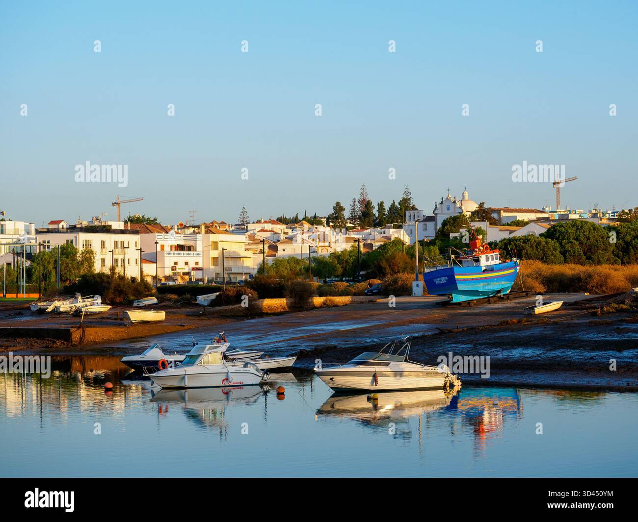 Ruhige Szene auf dem Fluss Gilao mit kleinen Booten und hellblauen Booten auf einer Slipway - die Stadt liegt im Hintergrund und 2 Kraniche sitzen am Horizont Stockfoto