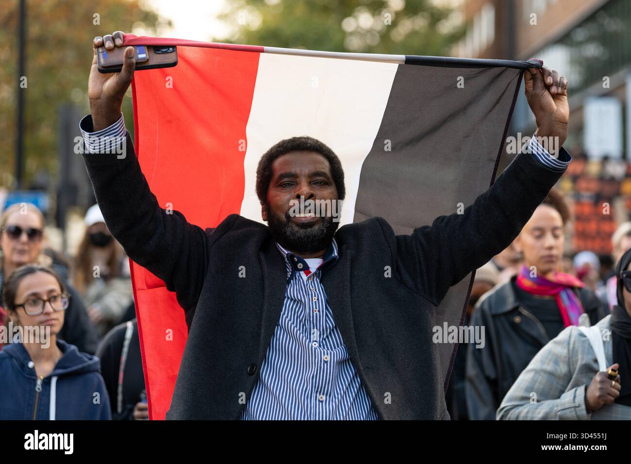 London, Großbritannien. November 2025. Ein Mann hält eine sudanesische Flagge, während er während der Kundgebung marschiert. Demonstranten marschierten in London, um Solidarität mit dem sudanesischen Volk zu zeigen und für ein Ende der britischen Waffenverkäufe an die VAE zu plädieren, die laut Aktivisten Gräueltaten der RSF-Miliz im ganzen Land ermöglichen. Quelle: SOPA Images Limited/Alamy Live News Stockfoto