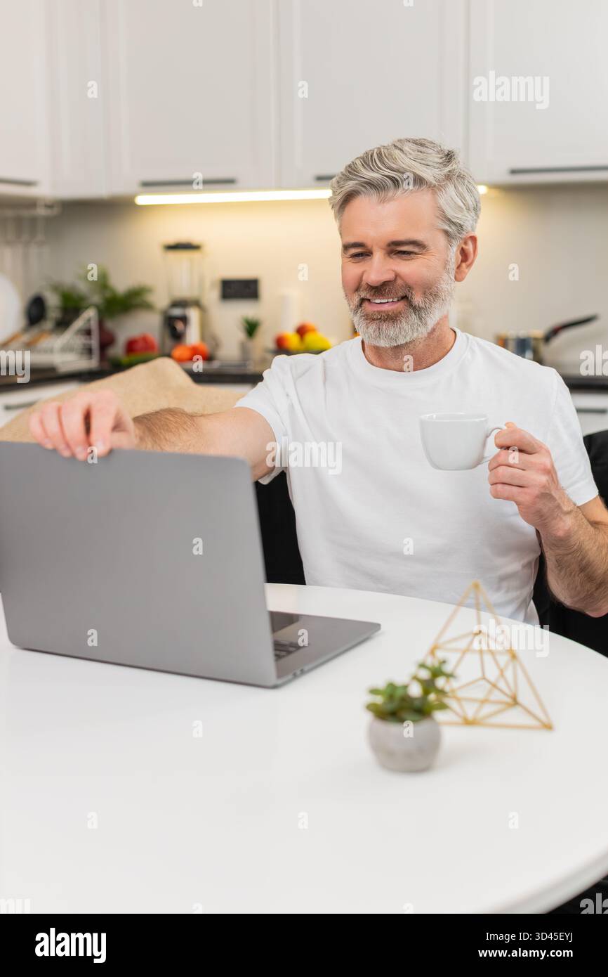 Ein Mann mittleren Alters zu Hause macht Espresso und öffnet den Laptop, um freiberuflich zu arbeiten. Ein reifer Mann, der am Tisch auf Sofas sitzt, lächelt mit einem Lächeln, plant Aufgaben und genießt eine ruhige Konzentration, bevor E-Mails frisch sind. Stockfoto