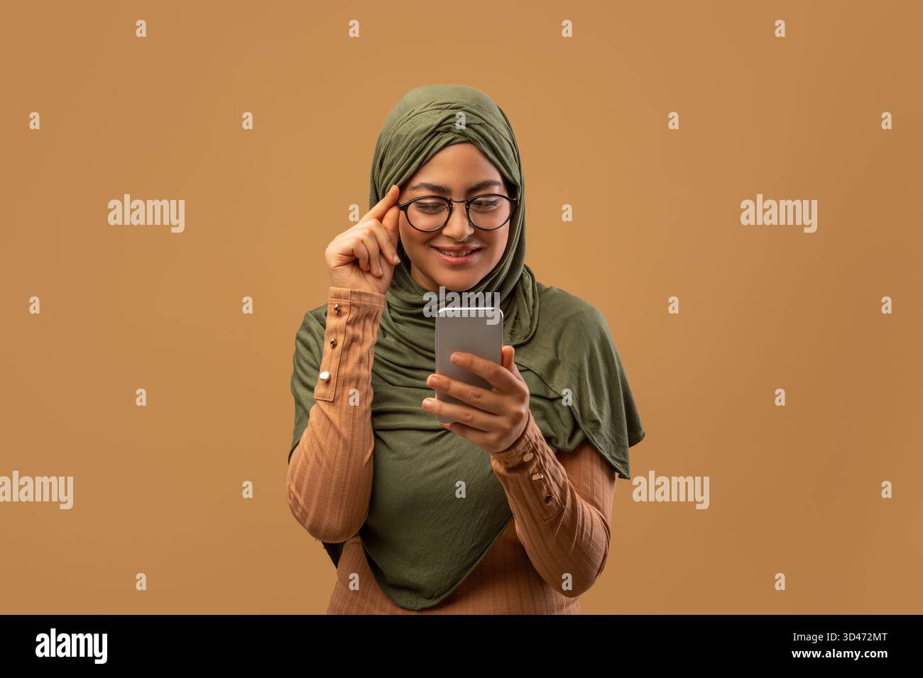 Positive muslimische Frau mit Kopftuch nutzt Smartphone, um neue App vor beigem Hintergrund zu erkunden Stockfoto