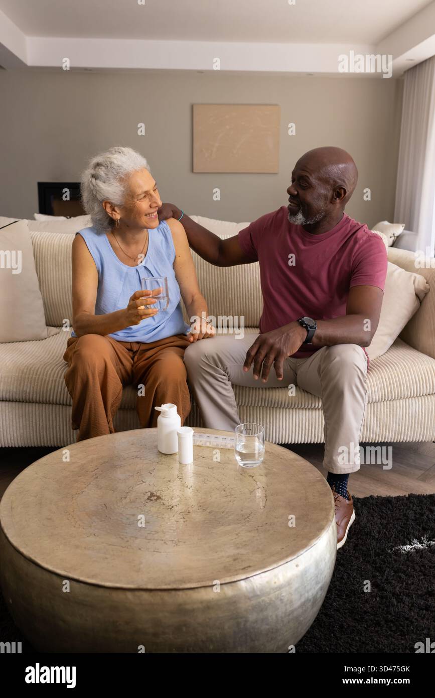 Diverse Seniorenpaare sitzen auf beigefarbener Couch im Wohnzimmer, mit Pillen-Organizer und Wasserglas Stockfoto