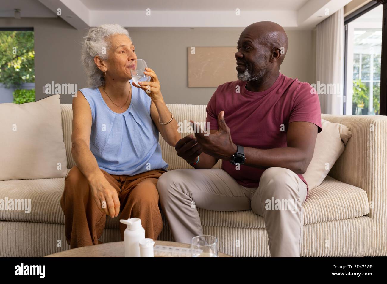 Trinken Sie Wasser und sprechen Seniorenpaar auf beigefarbenem Sofa im Wohnzimmer, mit Pillen-Organizer Stockfoto