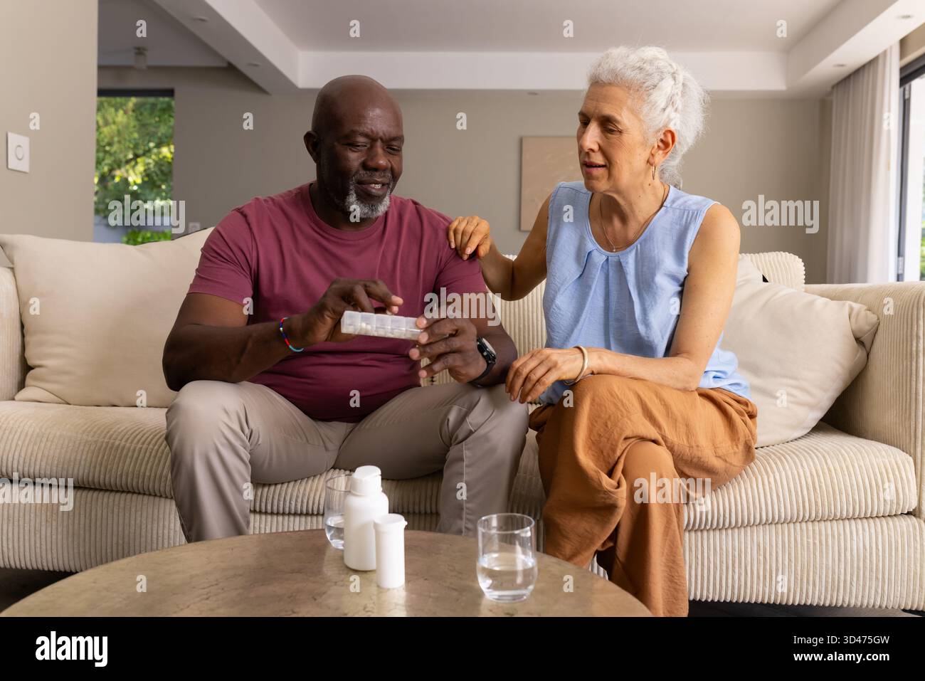 Verwaltung von Medikamenten diverse Seniorenpaare sitzen zu Hause auf Creme-Sofa, mit wöchentlichem Pillen-Organizer Stockfoto