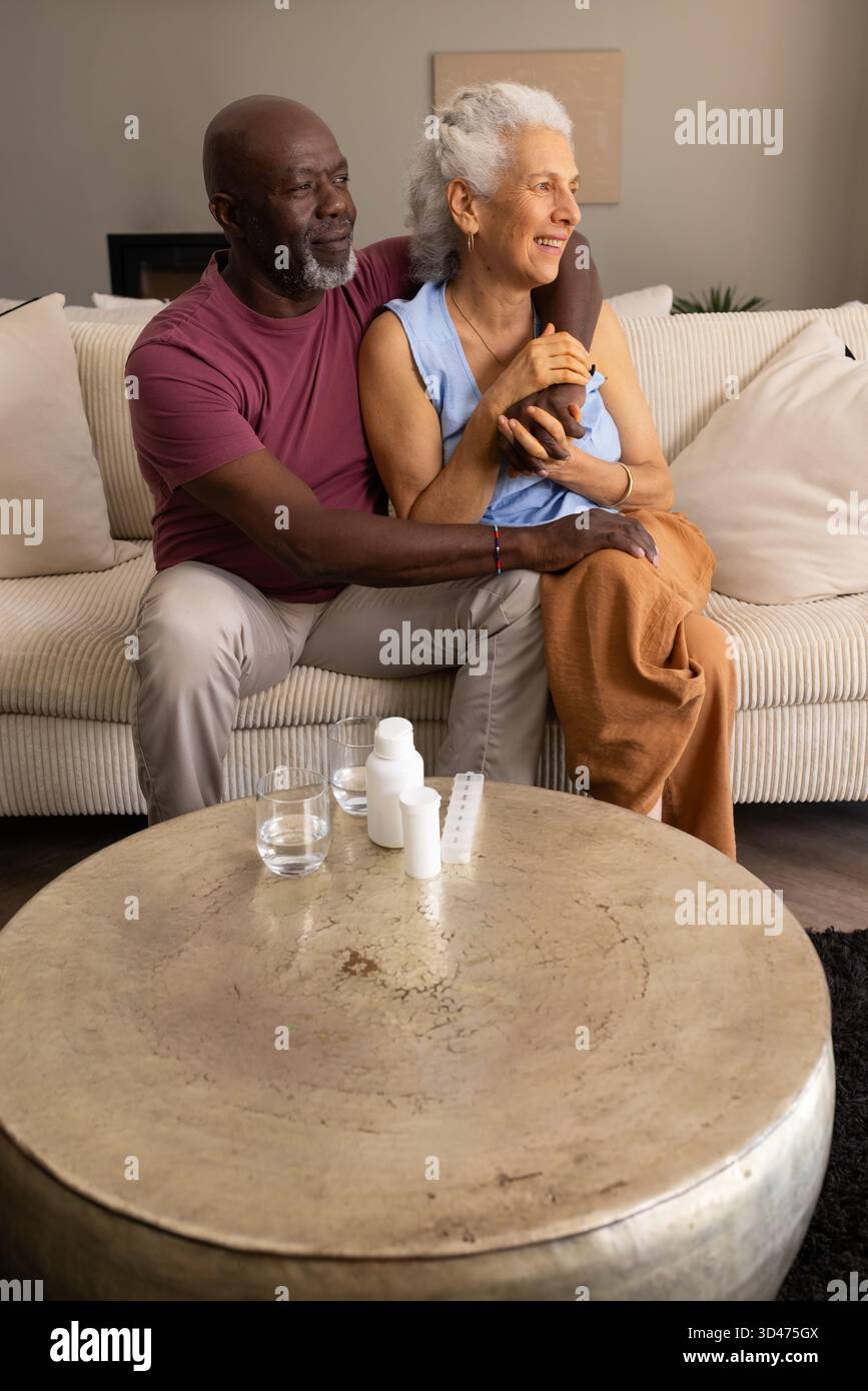 Lächelndes Seniorenpaar, das eng auf dem Sofa im Wohnzimmer sitzt, mit Pillendose und Medikamentenflaschen Stockfoto