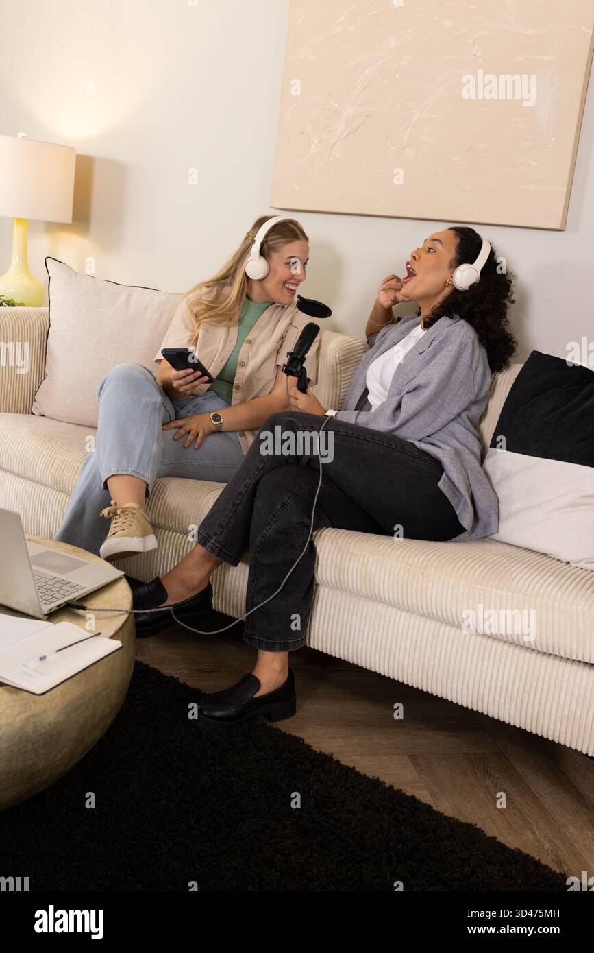 Verschiedene Freundinnen, die Podcasts im Heimstudio aufnehmen, mit schnurlosen Headsets und Laptop Stockfoto