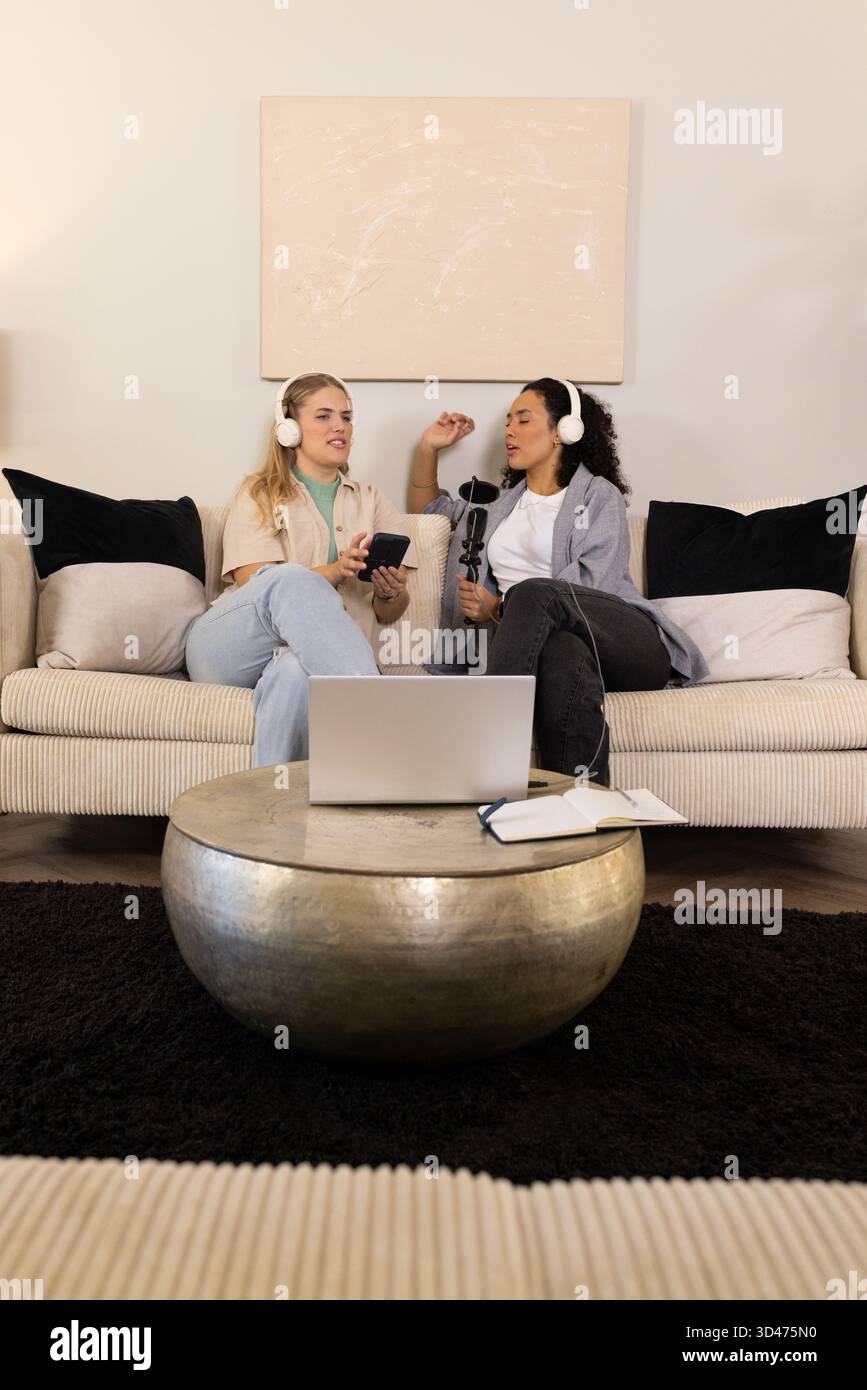 Verschiedene Freundinnen sitzen und Podcasts auf einem Wohnzimmer-Sofa mit Kopfhörern und Mikrofon aufnehmen Stockfoto