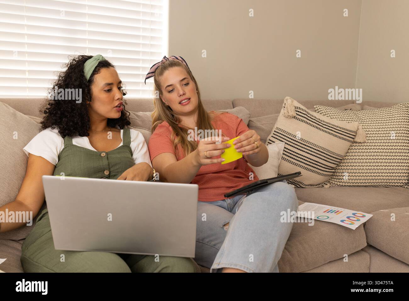 Zusammenarbeit mit verschiedenen Freundinnen, die auf einem beigefarbenen Sofa im Wohnzimmer sitzen, mit Laptop und Karten Stockfoto