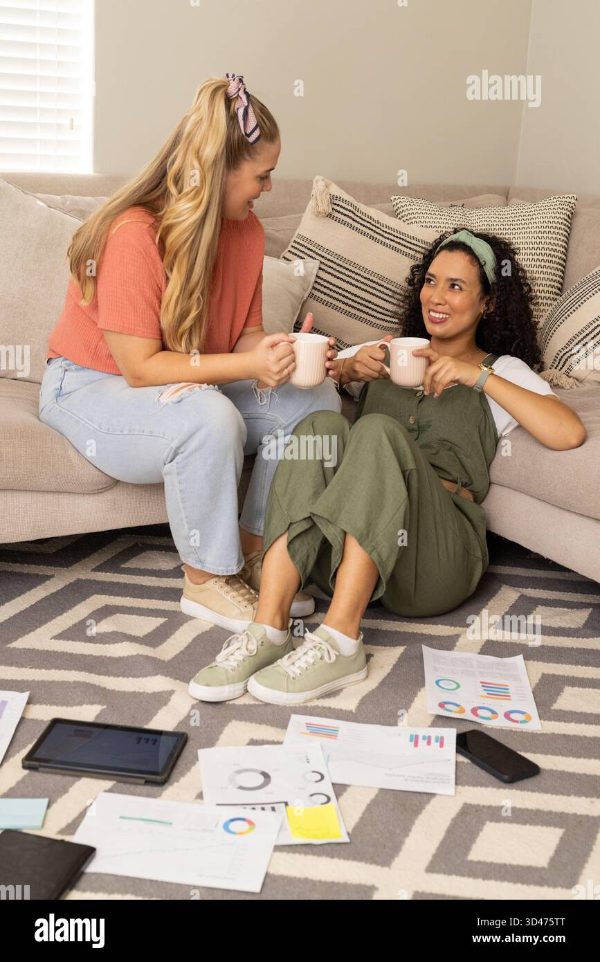 Sitzende Freundinnen, die Karten untersuchen und aus Keramikbechern im Wohnzimmer trinken, mit Tablet Stockfoto