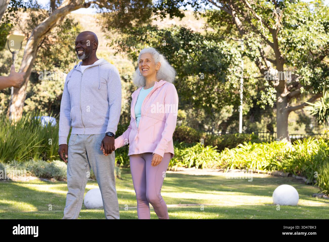 Spazieren Sie durch verschiedene Seniorenpaare, die Hände auf dem grasbewachsenen Parkrasen halten, mit Fitness-Tracker-Uhr Stockfoto