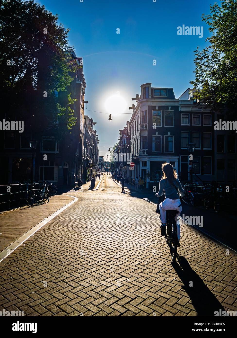 Eine junge Frau fährt mit dem Fahrrad auf einer Straße in Richtung Sonne in Amsterdam - Smartphone-aufgenommenes Stockfoto