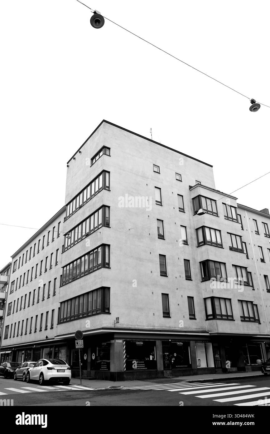 Eckansicht eines Wohnblocks mit Streifenfenstern, Autos und Straßenbahnleitungen, fotografiert in Helsinki, Finnland, in Schwarz-weiß an einem bewölkten Tag. Stockfoto
