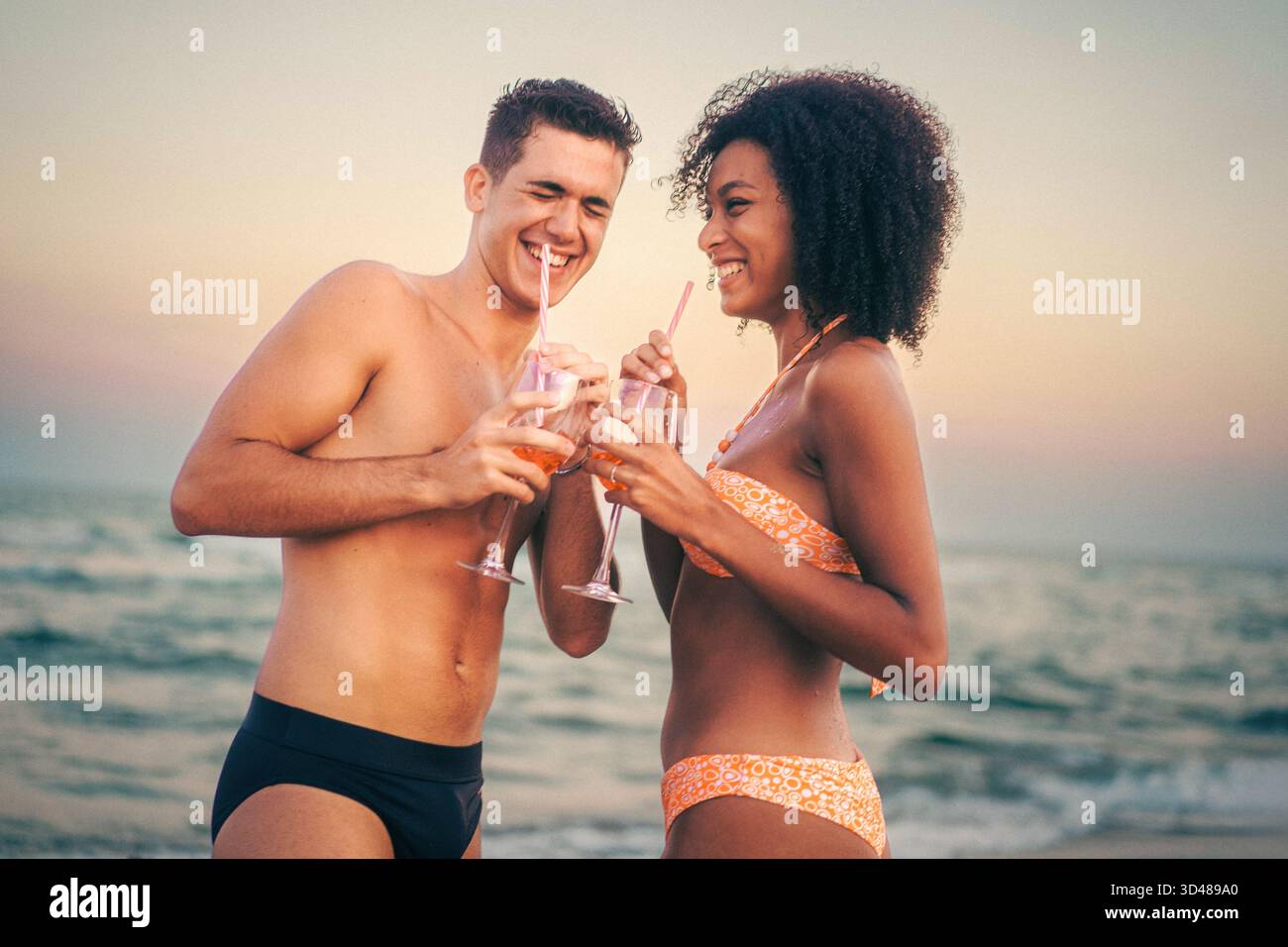 Ein junges Paar steht am Strand und schlürft bunte Cocktails durch Strohhalme. Sie teilen ein fröhliches Lächeln, wenn die Sonne untergeht, was eine warme und romantische Atmosphäre schafft Stockfoto