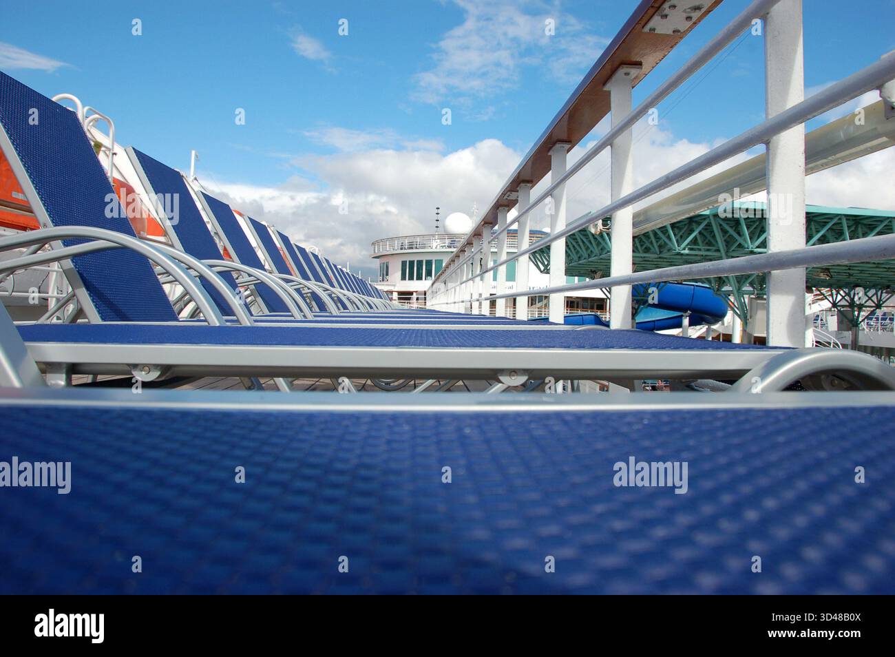 Reihe blauer Sonnenliegen auf einem Kreuzfahrtschiff unter hellem Himmel, aus einem niedrigen Winkel betrachtet, mit Geländern, Schiffsstrukturen und Wolken darüber Stockfoto