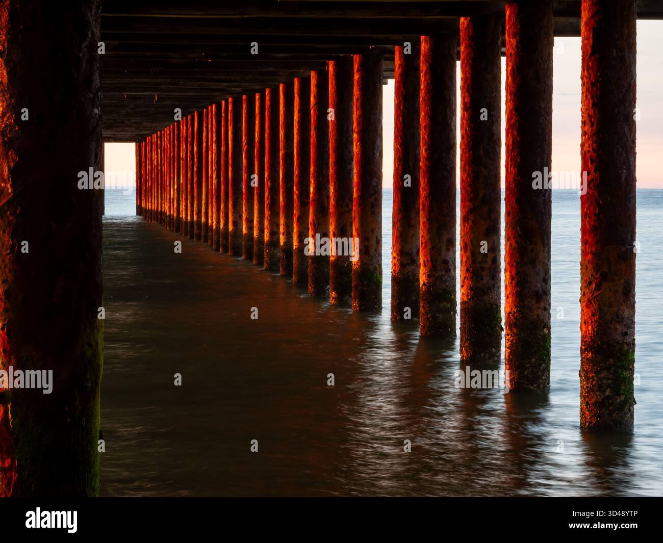 Unter dem Southwold Pier bei Sonnenaufgang mit warmem Licht, das die Stützpfeiler beleuchtet Stockfoto
