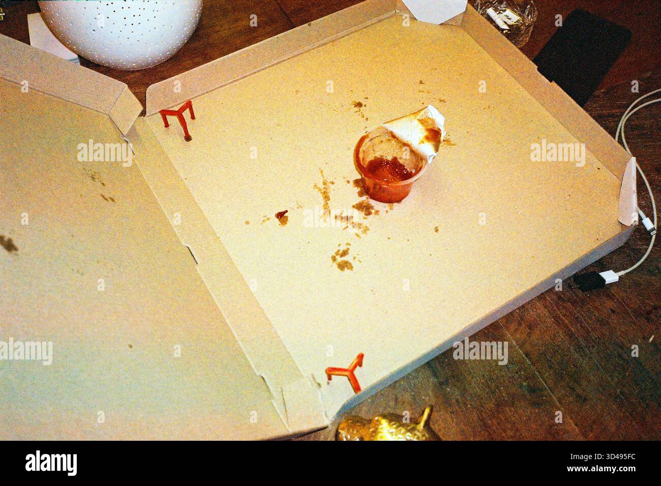 Leere Pizzaschachtel aus Pappe mit einer kleinen Tasse gebrauchten Ketchup, lässige, unordentliche Szene, analoger Film-Look mit alltäglichem Realismus. Stockfoto