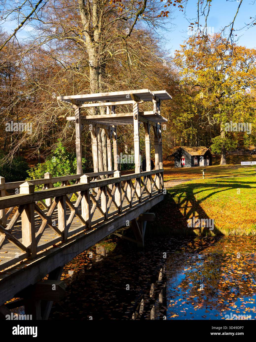 Holzbrücke über einen ruhigen Teich, umgeben von Herbstbäumen im warmen Sonnenlicht. Malerischer Blick auf ein Holzgebäude in einem friedlichen Park im Herbst. Stockfoto