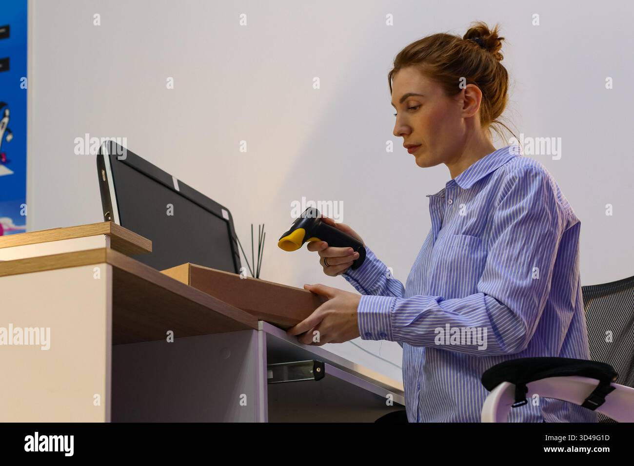 Mitarbeiter scannen kleine Pakete am Schreibtisch am Abholort. Junge Frau in gestreiftem Hemd hält Box und benutzt Barcode-Scanner am Schreibtisch mit Monitor. Stockfoto