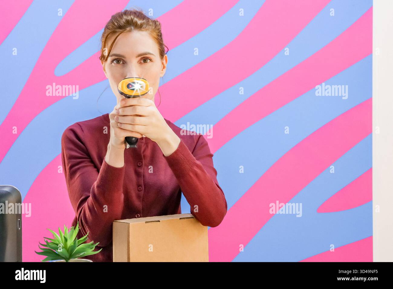 Frau, die Barcode-Scanner in der Nähe der Nase vor gestreiftem Hintergrund hält, mit Box auf dem Schreibtisch. Mitarbeiter am Abholort scannt spielerisch das Gesicht mit Barcode Stockfoto