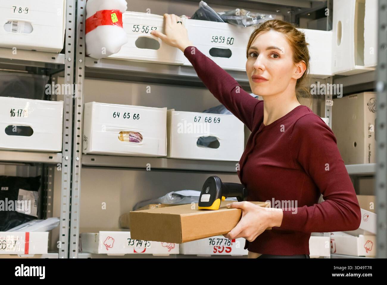 Weibliche Mitarbeiterin, die auf das Regal zeigt, während Box und Scanner im Aufbewahrungsbereich gehalten werden. Eine Frau in kastanienbraunem Hemd zeigt auf eine beschriftete Mülleimer in Metallregalen Stockfoto