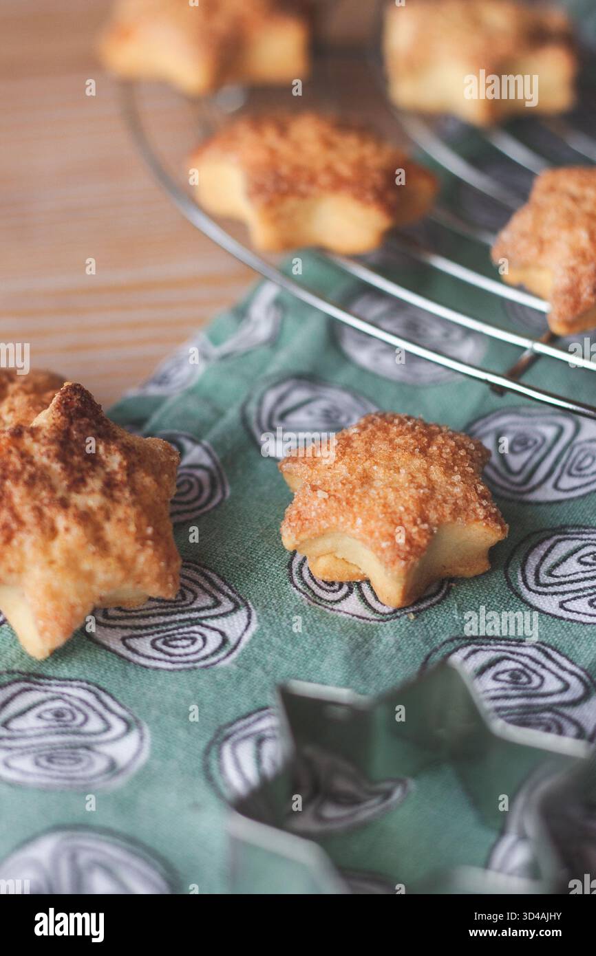 Nahaufnahme von hausgemachten Zimt-Sternkeksen, die auf einem Drahtgestell abkühlen. Warme, gemütliche Backatmosphäre mit gemustertem Küchentuch und weichem Tageslicht. Stockfoto