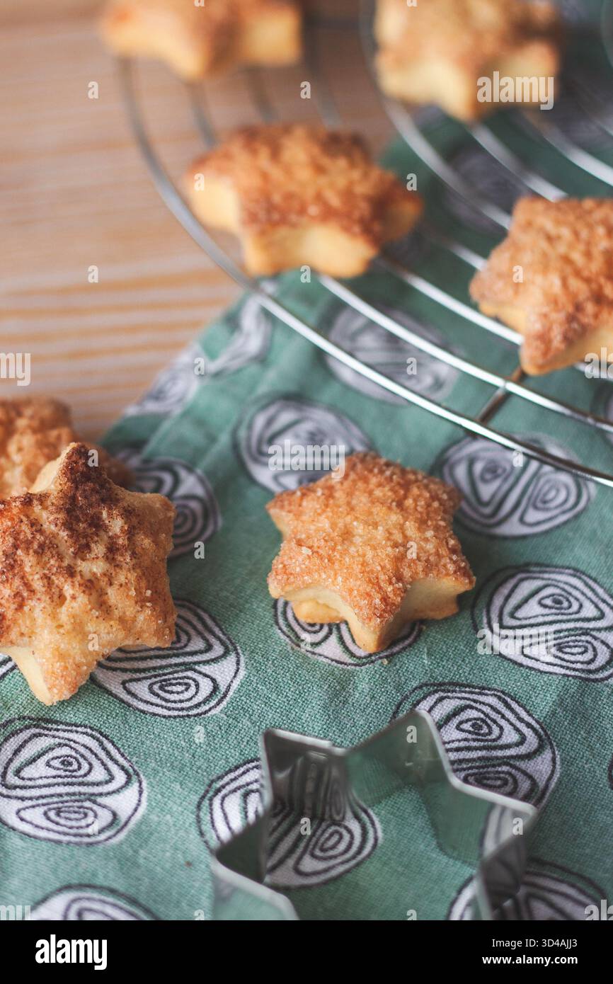 Frisch gebackene Zimt-Sternkekse auf einem Kühlregal, mit einem Metall-Stern-Ausstecher im Vordergrund. Gemütliche hausgemachte Weihnachtsbacken. Stockfoto