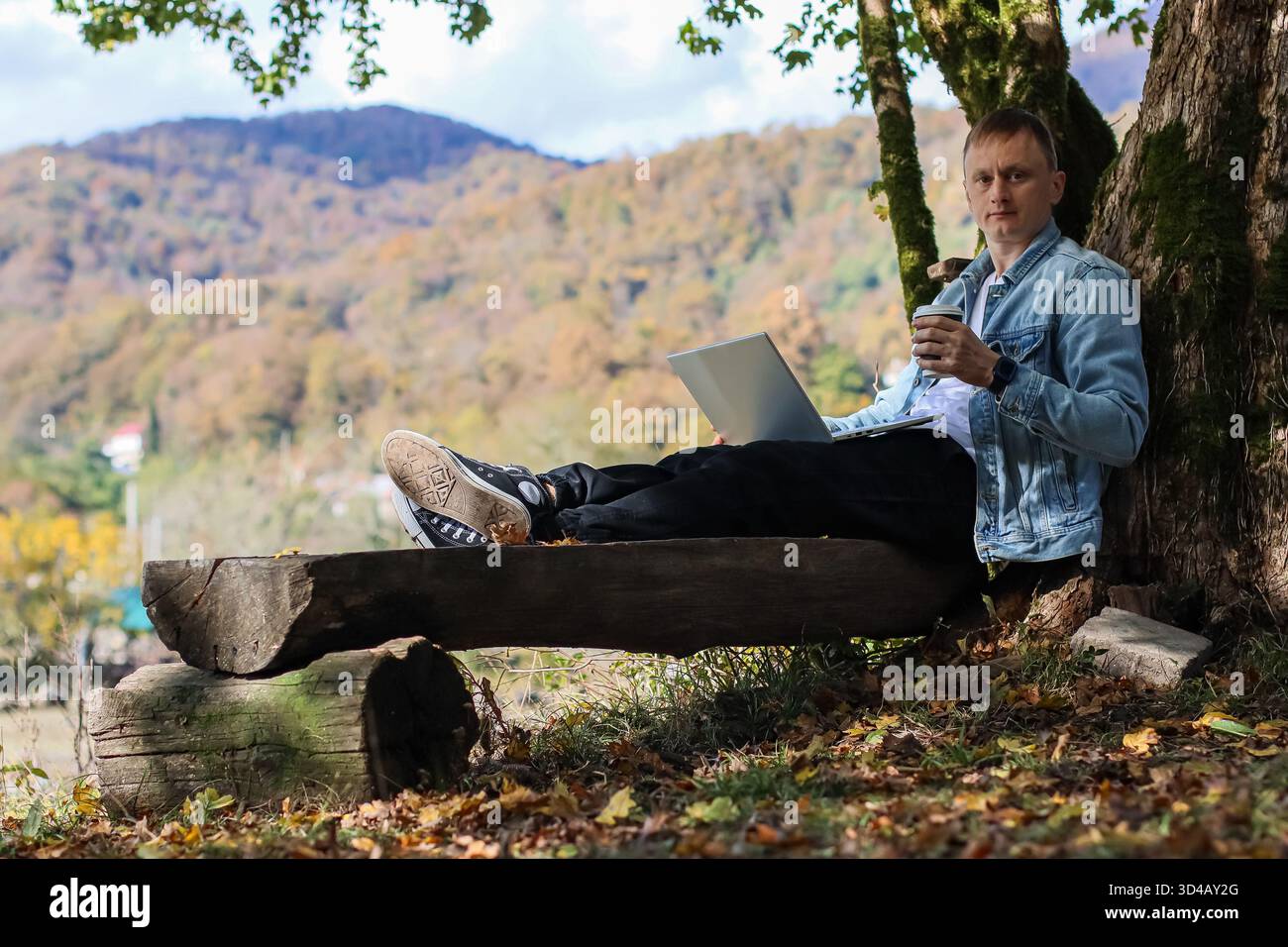 Mann balanciert Kaffee und arbeitet auf Laptop während Herbstwanderung. Mit erhobenen Füßen auf einem Holzstamm hält ein Mann während der Arbeit eine Kaffeetasse in einer Hand Stockfoto