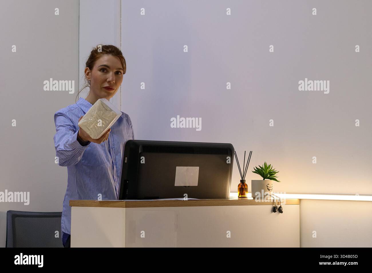 Frau, die versiegeltes Paket am Serviceschalter präsentiert. Der Mitarbeiter verstaut verpackte Artikel über den Schreibtisch mit Computer und kleinen Pflanzen Stockfoto