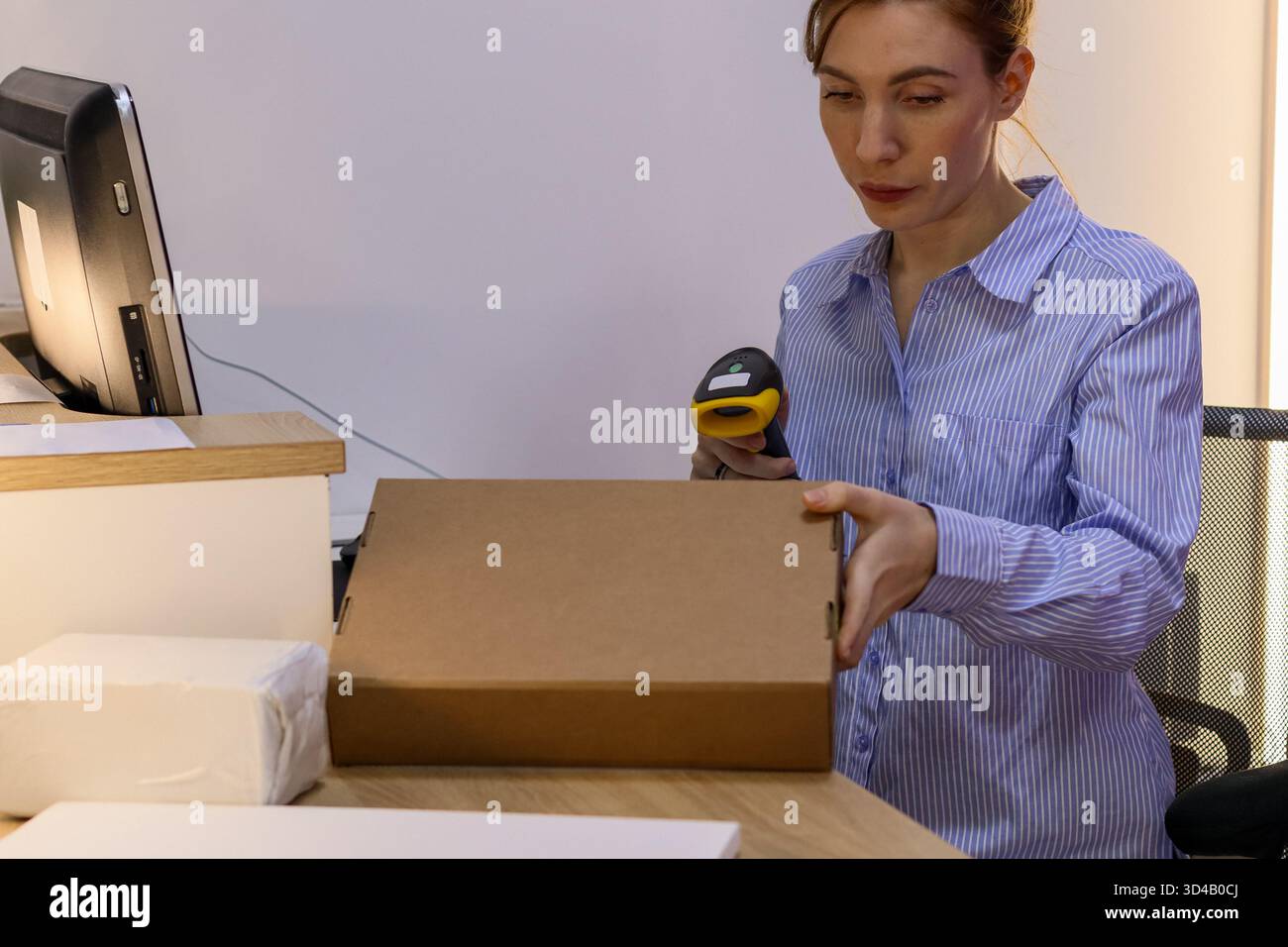 Frau, die braunen Pappkarton an der Theke scannt. Der Mitarbeiter hält eine flache braune Box, während er den Scanner in der Nähe des Computers im Abnahmebereich ausrichtet. Stockfoto