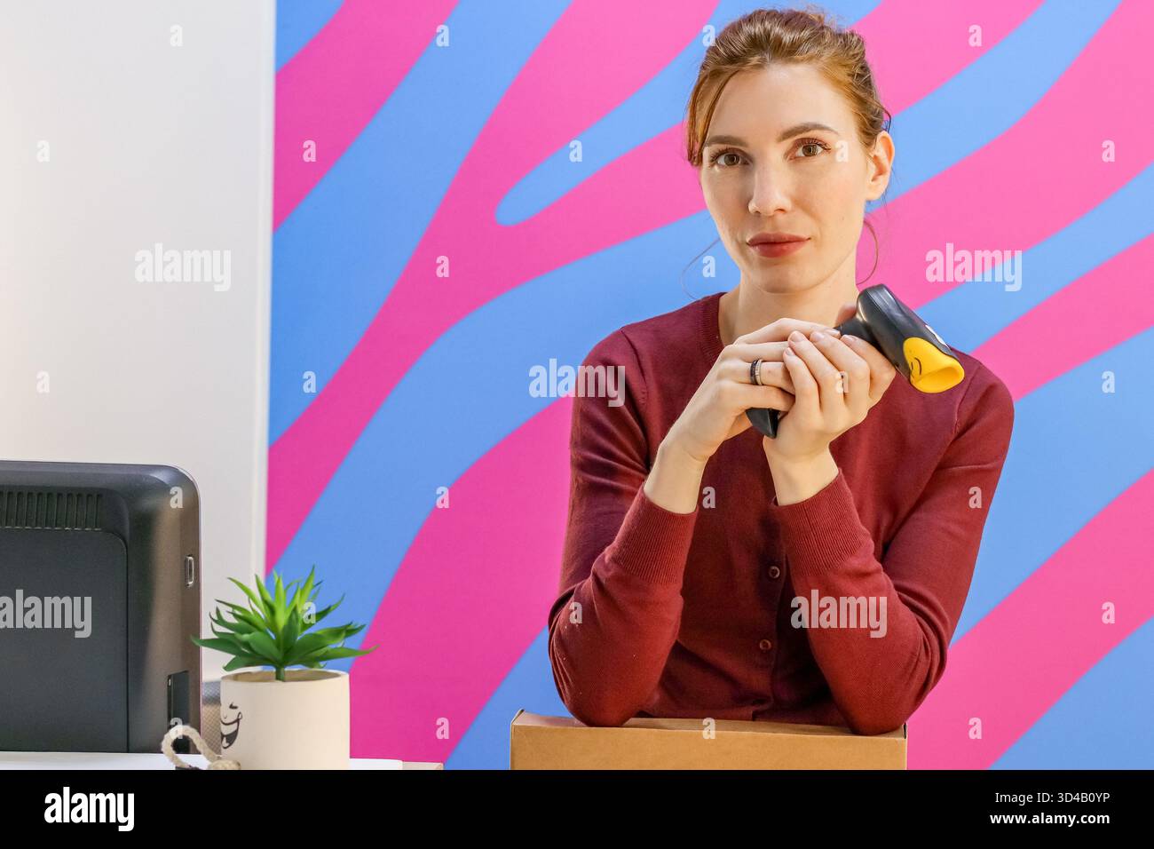 Frau zeigt Barcode-Scanner an der Abholstation. Mitarbeiter zeigt Handscanner in der Nähe des Schreibtisches mit Werk und Monitor Stockfoto