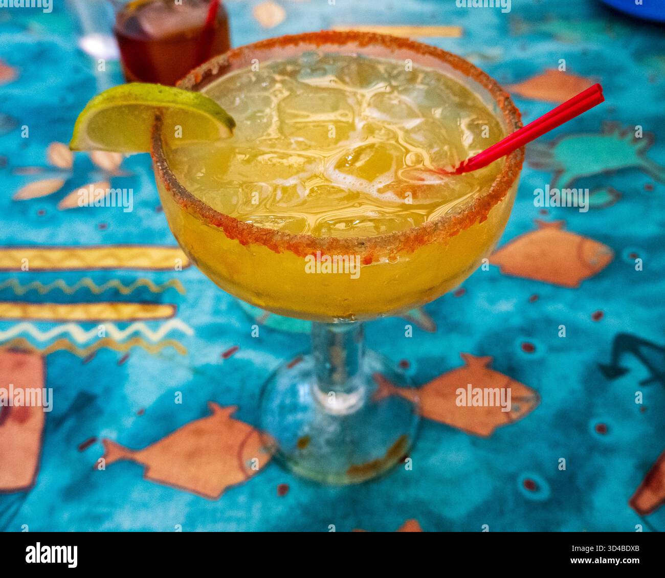 Eine erfrischende Margarita mit gesalzenem Rand und einer Zierleiste aus Limettenkeil sitzt auf einer leuchtend blauen Tischdecke mit orangefarbenen Fischen und geometrischen Mustern. Das Getränk Stockfoto