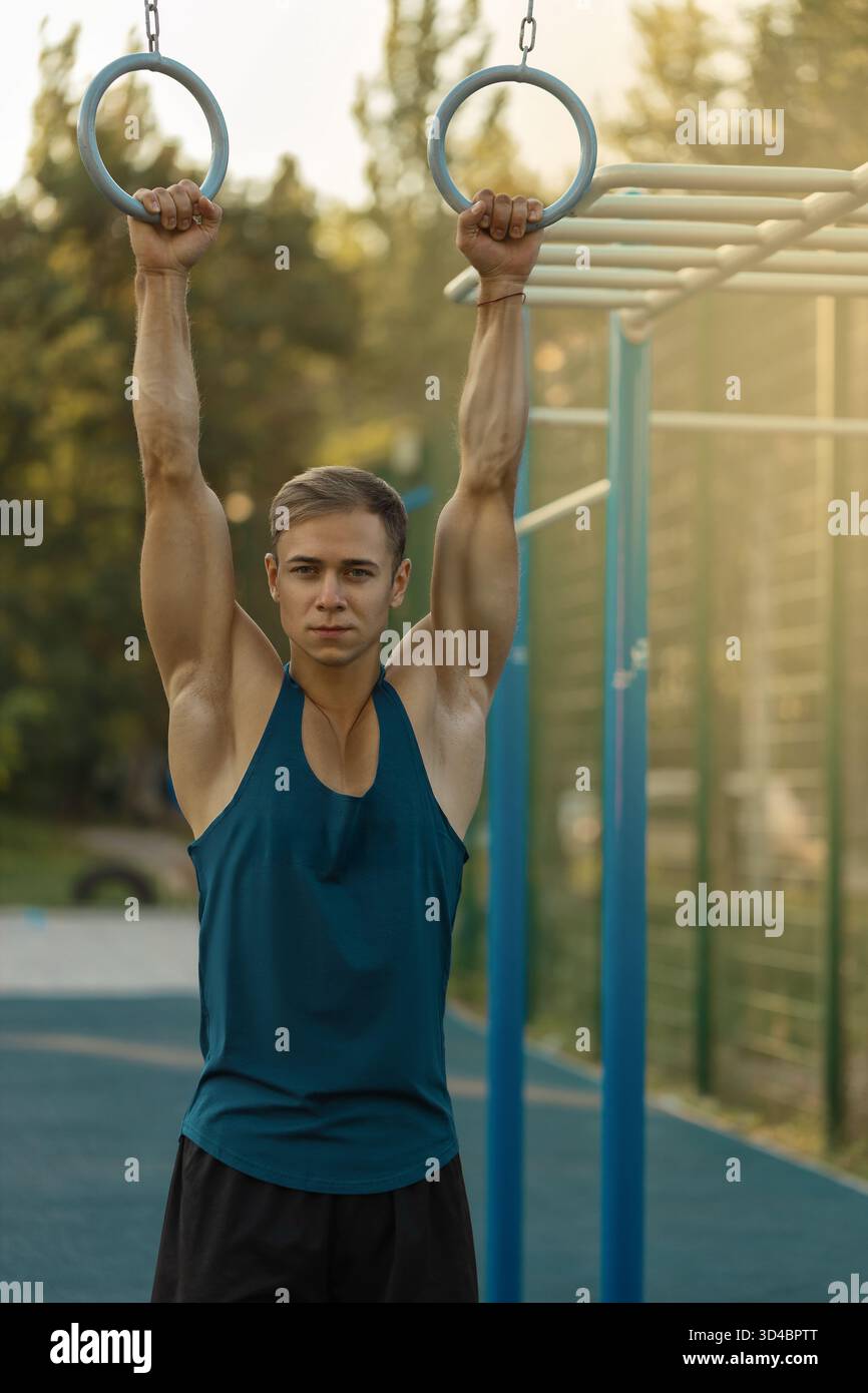 Männliche Athleten sind im Krafttraining aktiv und greifen Gymnastikringe auf einem Sportplatz. Er zeigt Entschlossenheit, während das Sonnenlicht seinen konzentrierten Ausdruck und seine Muskeln hervorhebt. Stockfoto