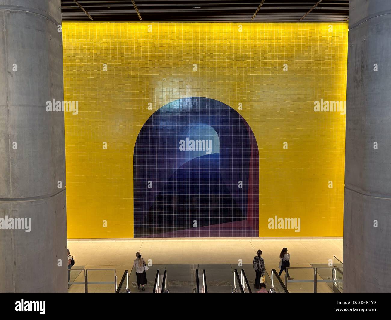 Leuchtend gelb geflieste Wand mit dunkelblauem Bogengemälde in einer modernen U-Bahn-Station in Sydney. Pendler auf Rolltreppen unter dem öffentlichen Kunstwerk. Stockfoto