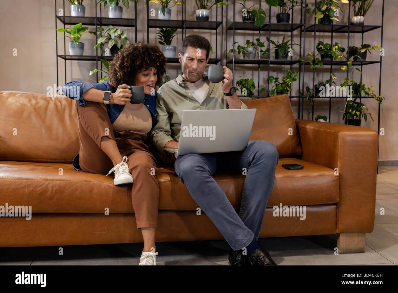 Stöbern Sie durch verschiedene Paare, die einen Laptop auf brauner Ledercouch im Wohnzimmer mit Kaffeetassen bewerten Stockfoto