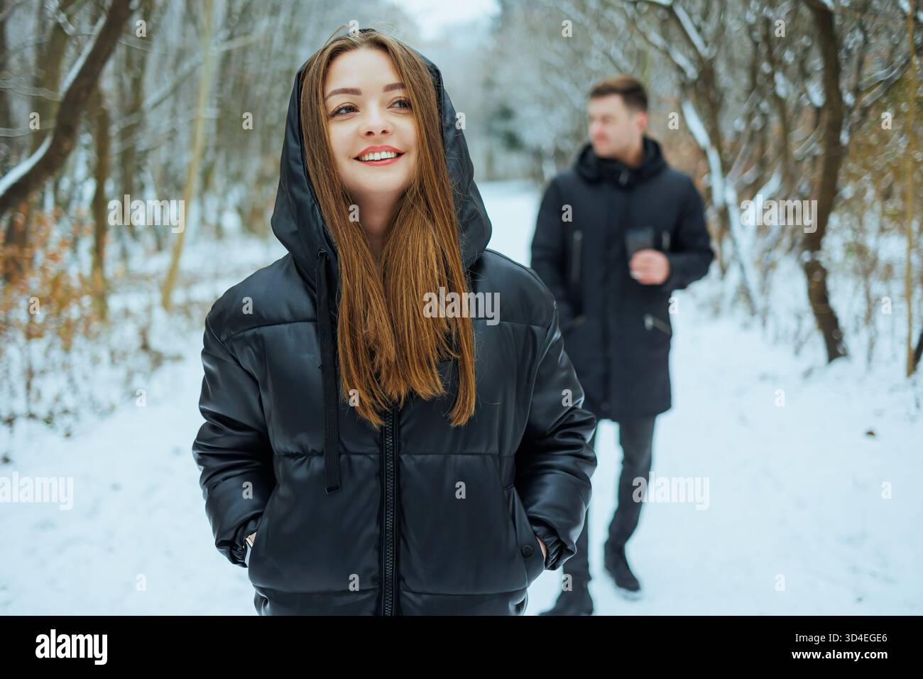 An einem fröhlichen Wintertag spaziert eine fröhliche junge Frau in einem stilvollen schwarzen Mantel auf einem verschneiten Pfad, während ihr Begleiter hinter ihr schlendert Stockfoto