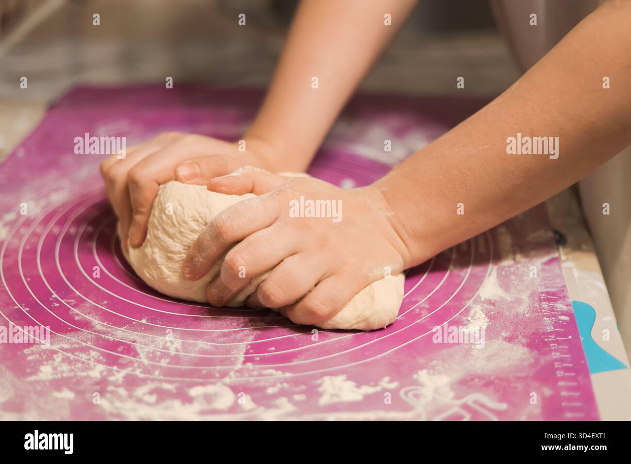 Eine Nahaufnahme von Händen, die einen hellen Teig fest auf einer rosa oder violetten Silikon-Backmatte kneten. Hochwertige Fotos Stockfoto