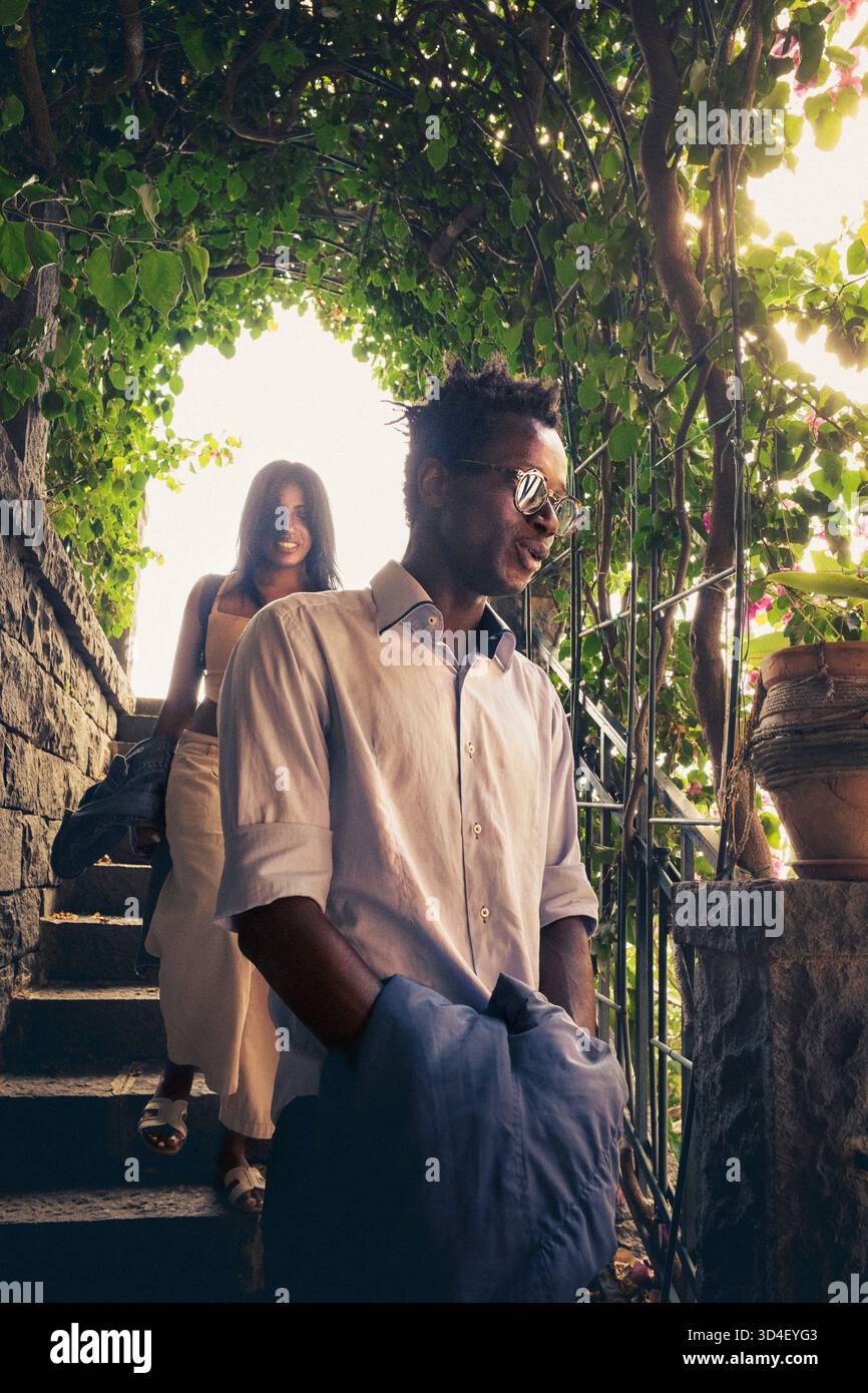 Ein charmantes Paar geht Hand in Hand entlang einer Steintreppe, die von üppigen grünen Weinstöcken beschattet wird. Die Sonne filtert durch die Blätter und erzeugt ein warmes und römisches Licht Stockfoto
