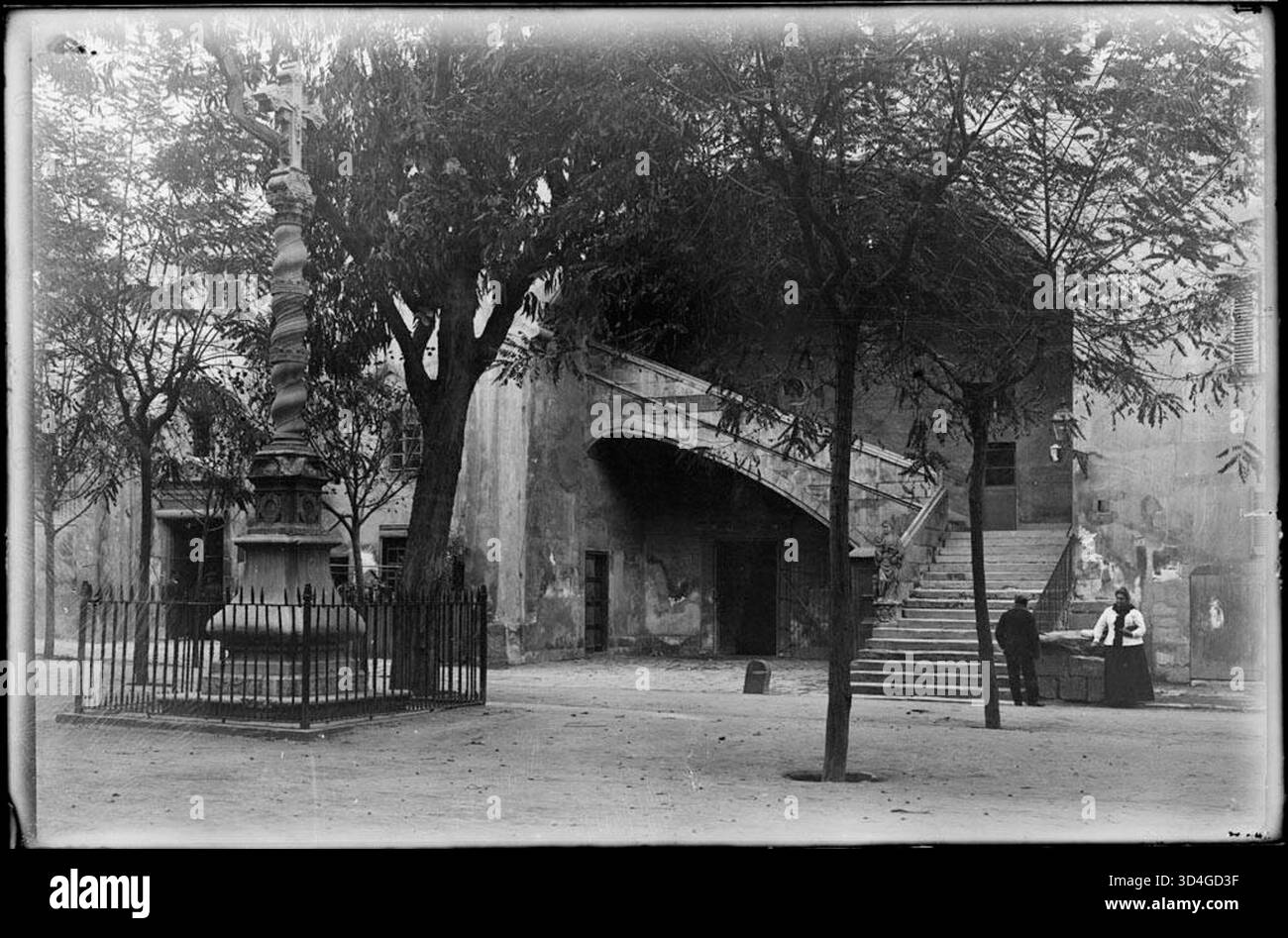Restaurierte Fotografie von Josep Maria Armengol i Bas aus dem Innenhof des Hospital de la Santa Creu, mit einem Kreuz in der Mitte und zwei Personen in der Nähe der Eingangstreppe, aufgenommen zwischen 1899 und 1915. Stockfoto