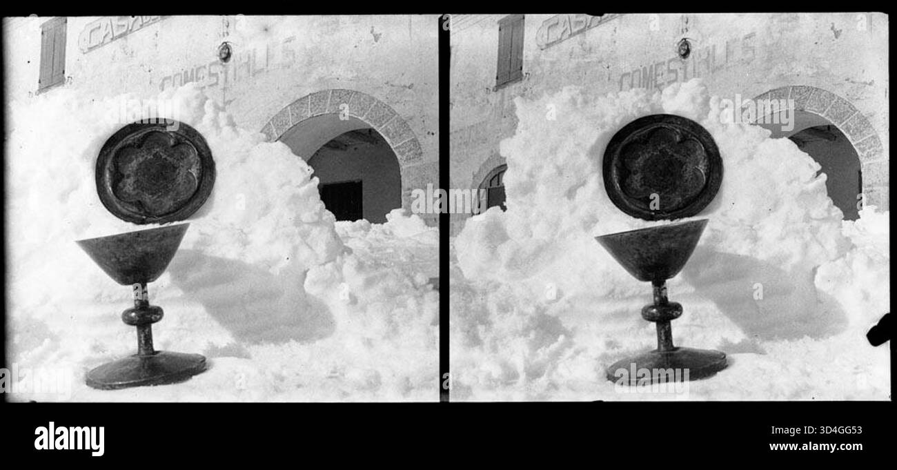 Ein Kelch und ein weiteres Metallobjekt auf einem Schneehaufen vor einem Gebäude mit einem Schild mit der Aufschrift „Comestibles“ in Núria, das von Josep Domènech i Sàbat zwischen 1922 und 1934 gefangen genommen wurde. Das Bild zeigt eine Winterszene mit historischem Kontext. Stockfoto