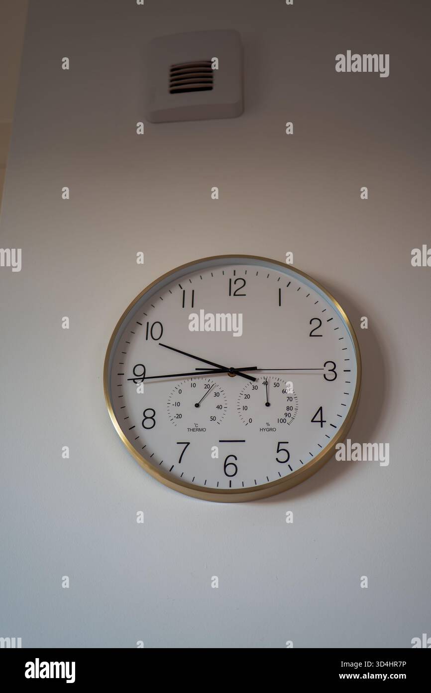 Eine rund-um-Uhr mit goldenem Rand, schwarzen Ziffern sowie Temperatur- und Feuchtigkeitsregler hängt an einer hellen Wand unter einem kleinen quadratischen Lüftungsschlitz in einem hellen, modernen Apartment Stockfoto