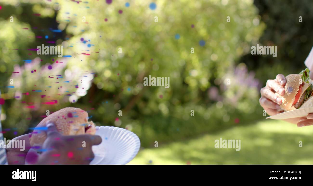 Zwei Männer mit kurzen Ärmeln, die Burger mit Salat auf Papiertellern im sonnigen Garten halten, Konfetti Stockfoto