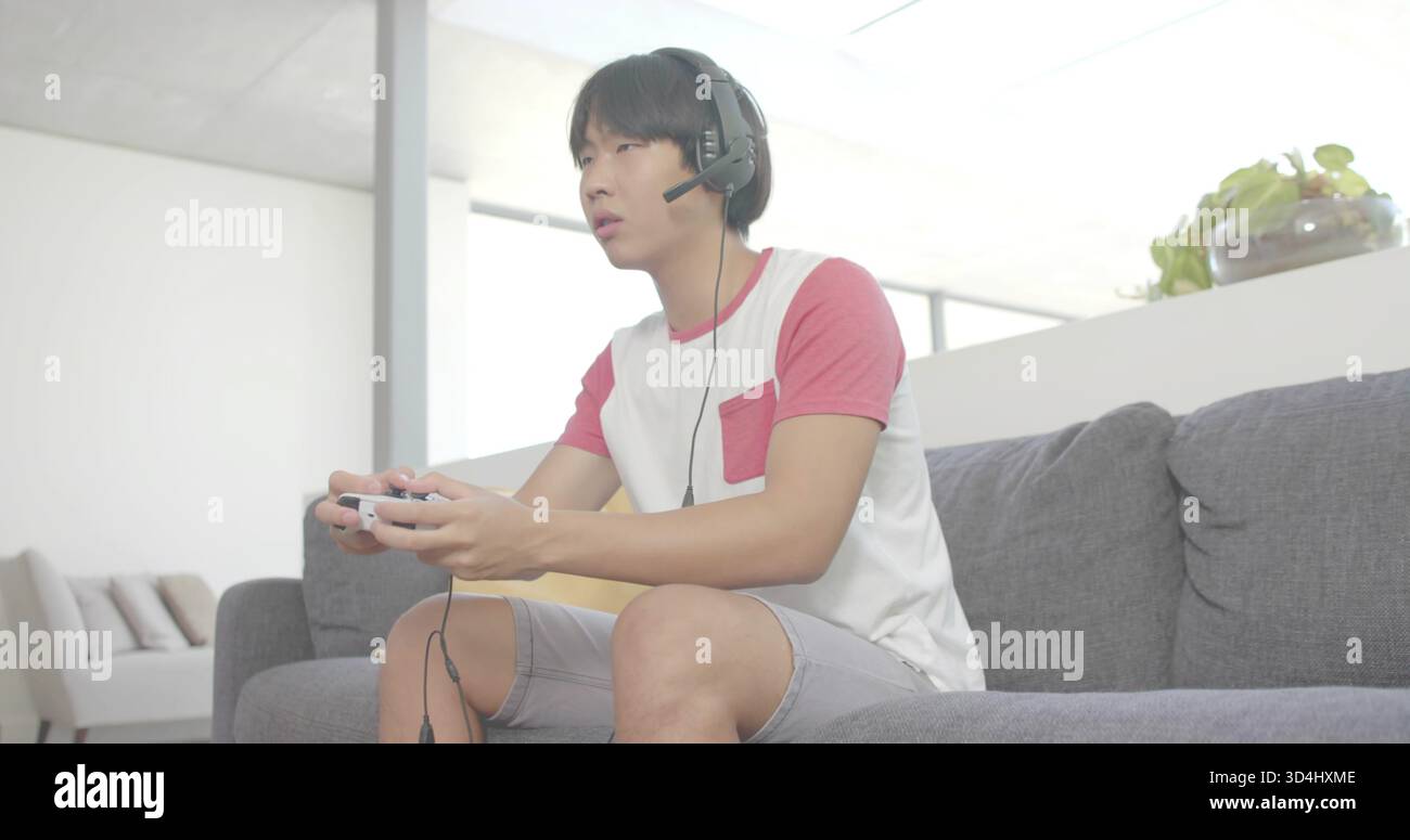 Spielender Mann mit weißen T-Stück roten Ärmeln, der kabelgebundenen Controller auf grauem Sofa zu Hause hält, Headset Stockfoto