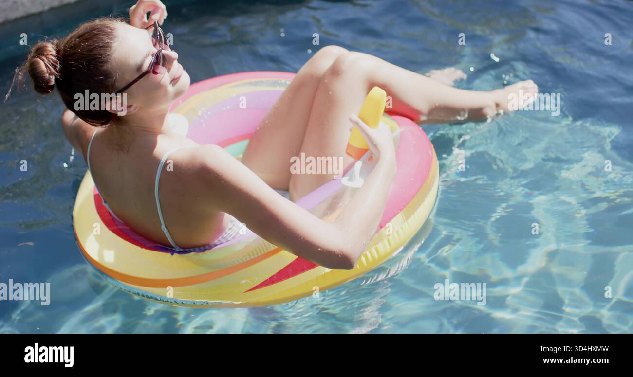Schwimmende Frau in Badeanzug und Sonnenbrille auf Regenbogenfloat mit Tasse mit Orangengetränk, Poolrand Stockfoto