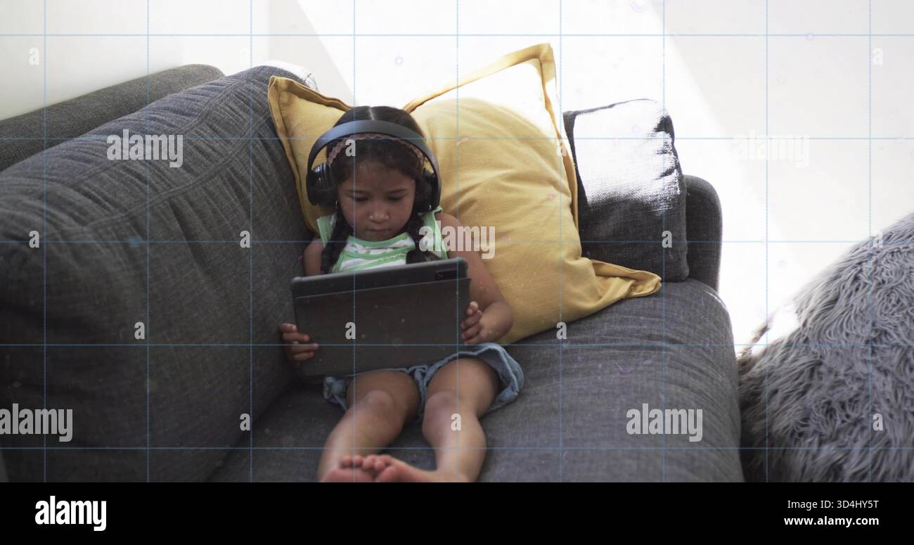 Liegendes Kind trägt Kopfhörershorts mit gestreiftem Oberteil, die ein Tablet auf einem grauen Sofa hält, Senfkissen Stockfoto