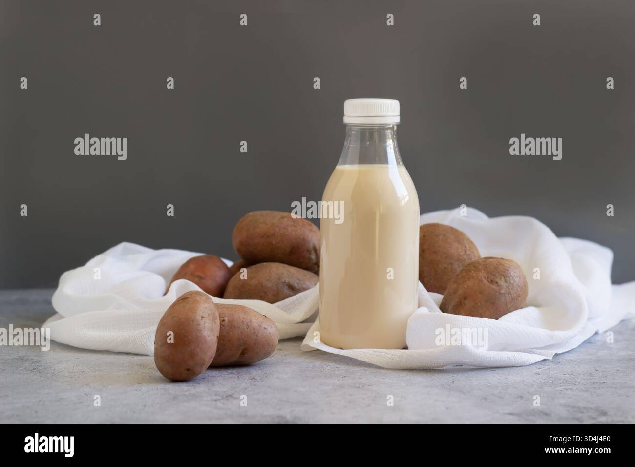 Flaschenmilch steht in der Mitte, umgeben von ganzen, ungeschälten Kartoffeln auf einem weißen Tuch. Die Komposition hebt das natürliche hervor Stockfoto