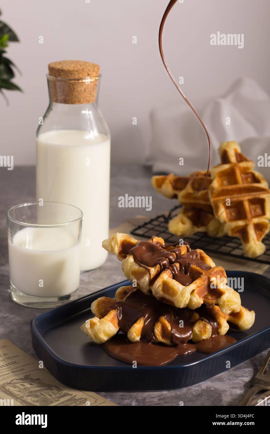 Waffeln auf einem Teller werden mit reichhaltiger Schokoladensauce getränkt, begleitet von einem Glas Milch und einer Flasche Milch in einer rustikalen Küche, cr Stockfoto