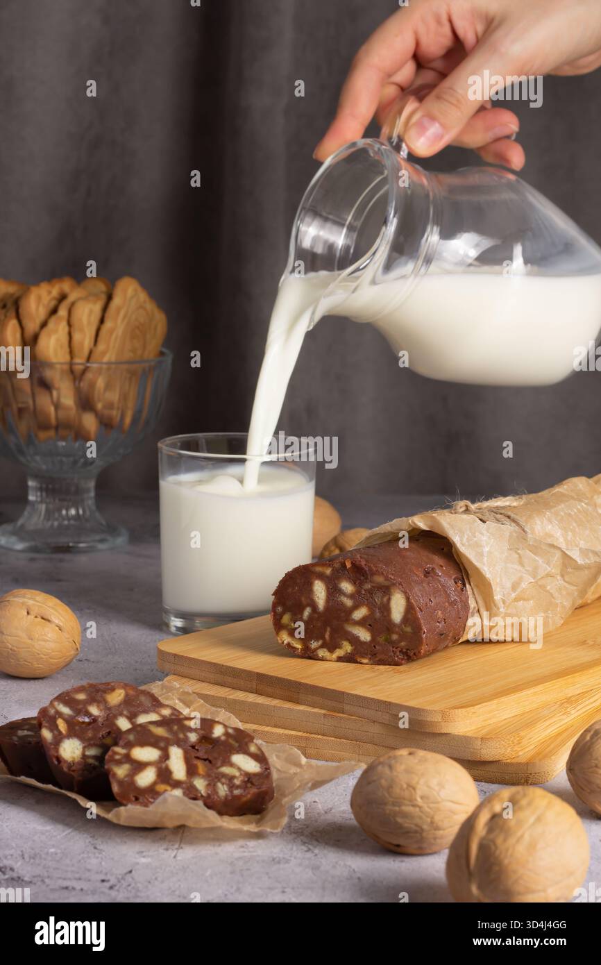 Eine Hand gießt frische Milch in ein Glas neben einer Platte mit Schokoladensalami und Keksen. In der gemütlichen Umgebung befinden sich Walnüsse im Vordergrund und ein Desse Stockfoto